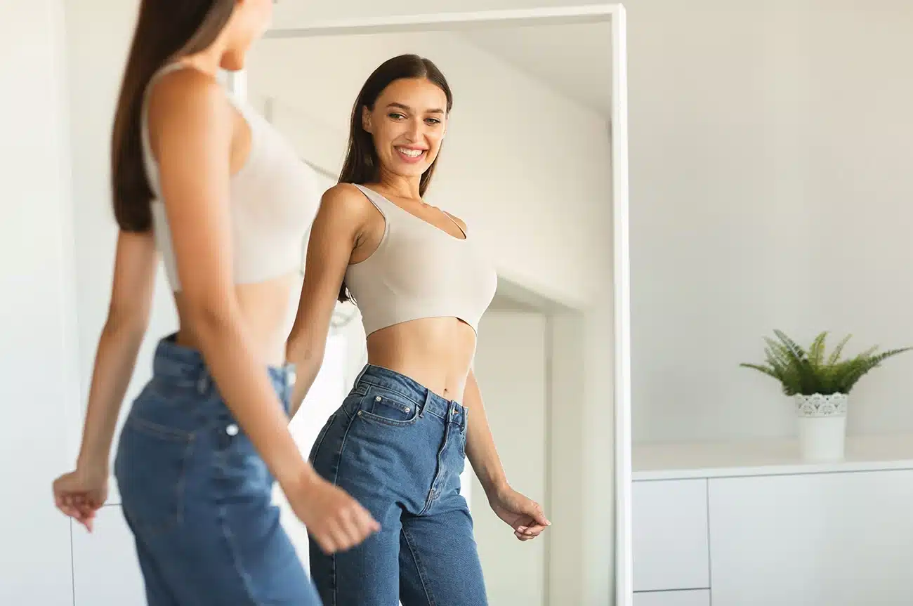 Smiling woman looking at her slim waist and reflection in a full-length mirror. Smiling woman looking at her slim waist and reflection in a full-length mirror.