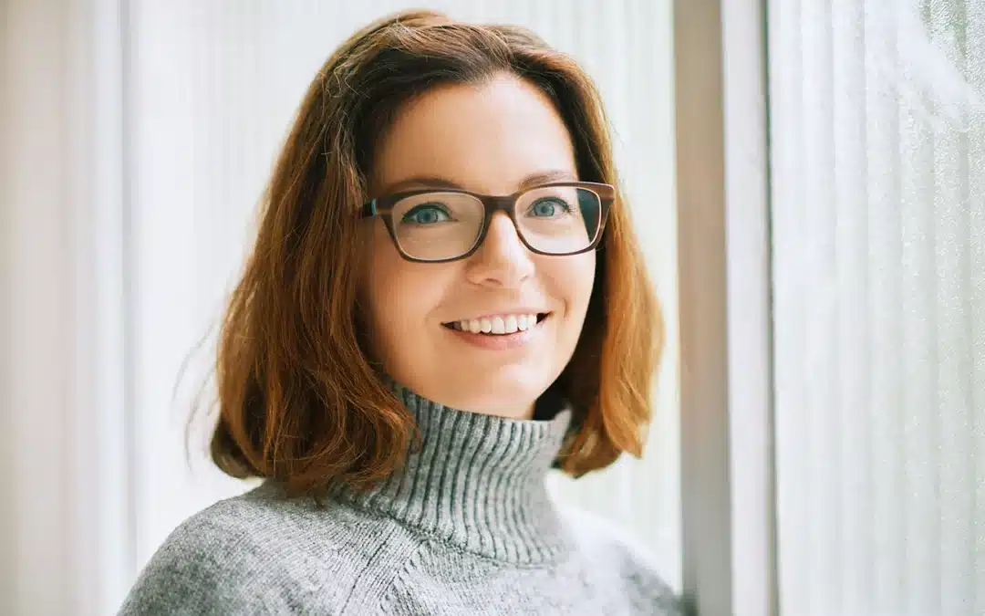 Smiling woman with brown hair and glasses leaning near a window indoors.